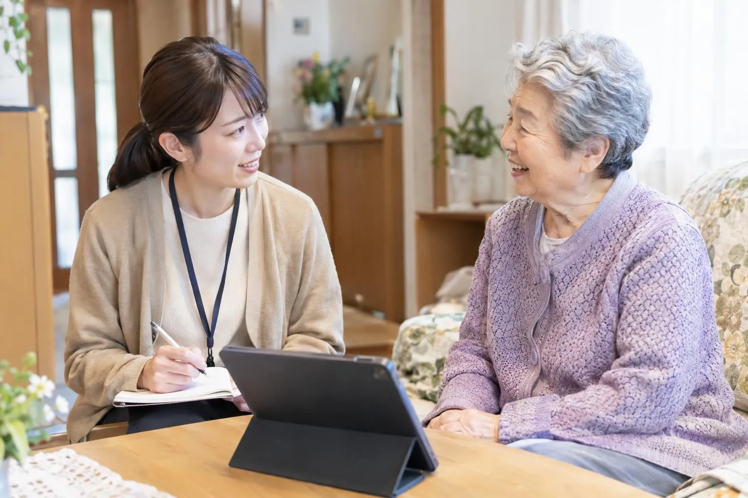 自宅を訪ねてタブレットの横で話を聞く担当者と高齢の女性